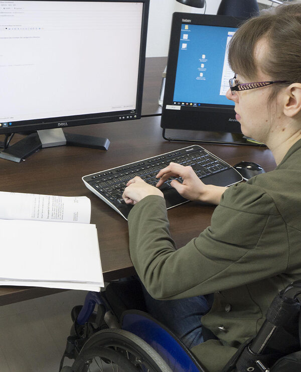 Disabled worker in a wheelchair typing at keyboard while looking at two computer screens.
