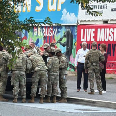 A group of uniformed soldiers gather around the back of a pickup truck on a city street, while a few civilians stand nearby. Behind them are colorful murals and a large sign advertising live music events.