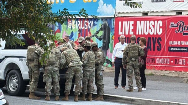 A group of uniformed soldiers gather around the back of a pickup truck on a city street, while a few civilians stand nearby. Behind them are colorful murals and a large sign advertising live music events.