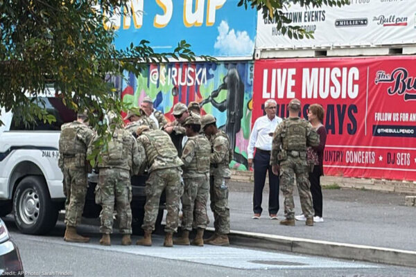 A group of uniformed soldiers gather around the back of a pickup truck on a city street, while a few civilians stand nearby. Behind them are colorful murals and a large sign advertising live music events.