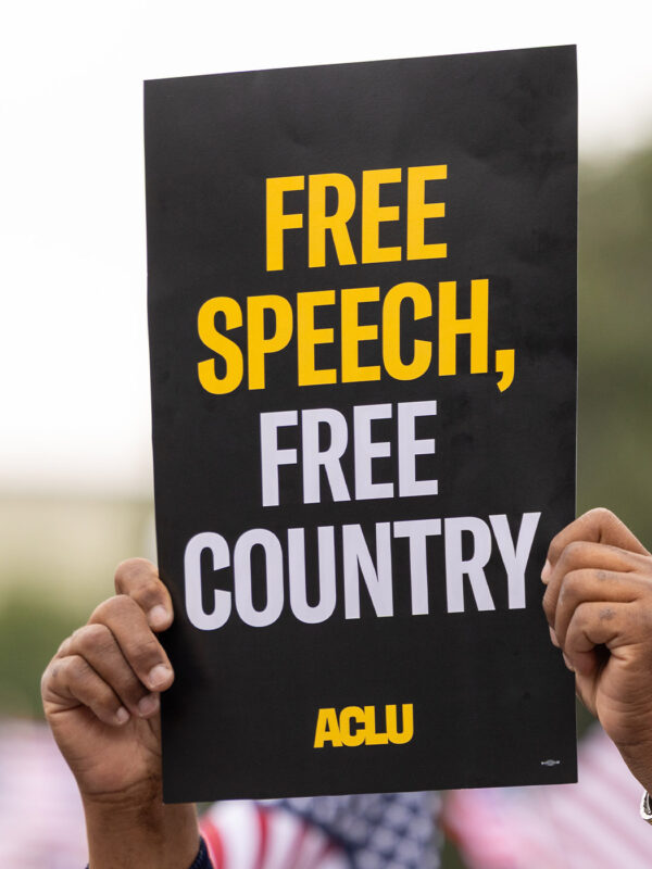 A demonstrator (with only their hands visible) holds up a sign that reads " FREE SPEECH, FREE COUNTRY - ACLU".