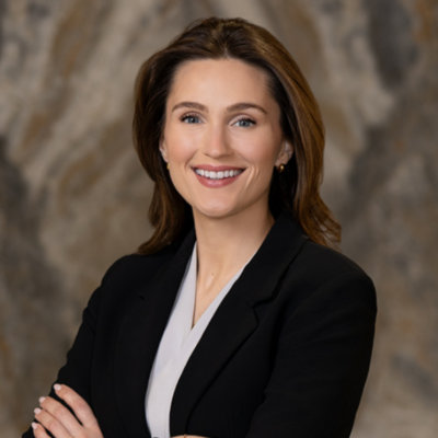 Pictured is Laura Follansbee, she has brown hair and blue eyes. Laura has her arms crossed and is wearing a dark blazer and a white blouse.