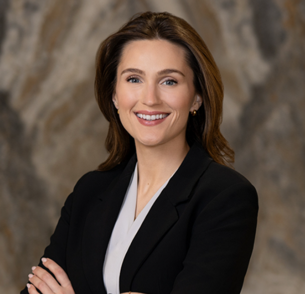 Pictured is Laura Follansbee, she has brown hair and blue eyes. Laura has her arms crossed and is wearing a dark blazer and a white blouse.