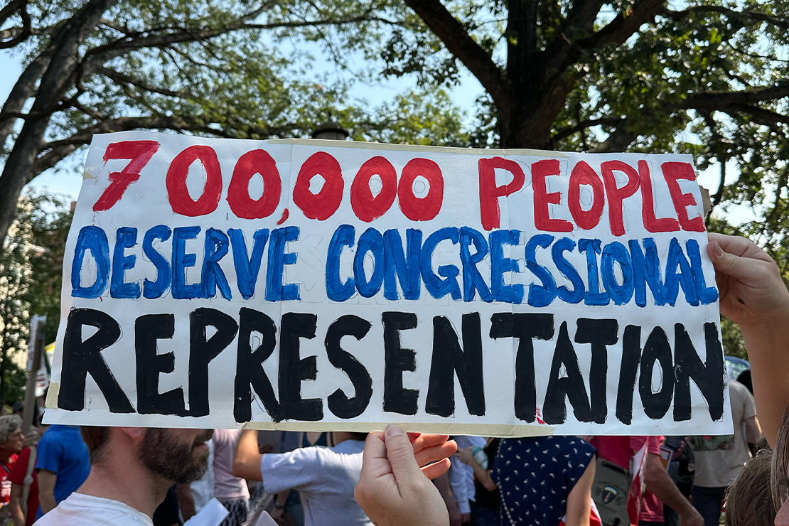 A person at a rally holds a handmade white sign with large red, blue, and black lettering that reads "700,000 People Deserve Congressional Representation," with a crowd gathered under trees in the background.