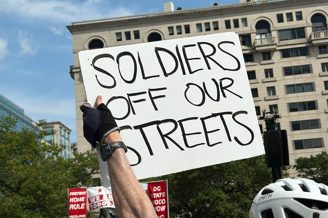 A protester holds a white handmade sign reading "Soldiers Off Our Streets" in large black letters during a DC statehood rally, with city buildings, trees, and red protest signs in the background.