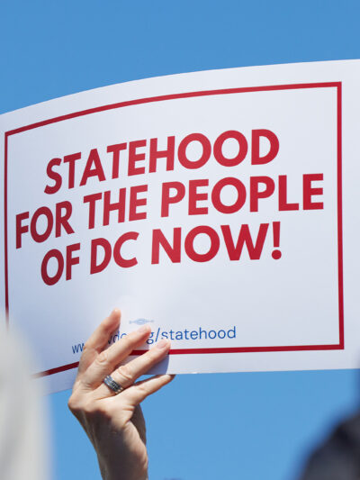 A person holds a white sign with bold red text and a red border reading "Statehood for the People of DC Now!" against a bright blue sky, with two blurred people in the foreground.