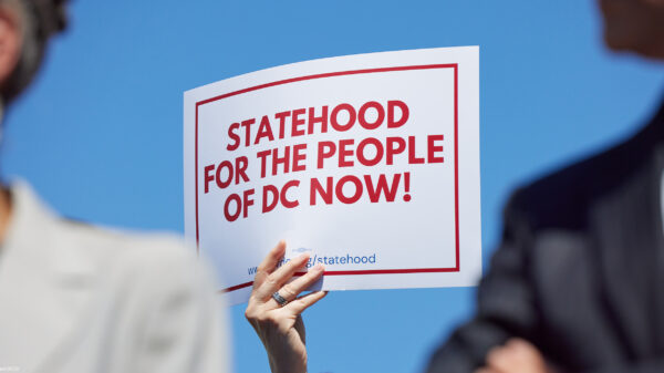 A person holds a white sign with bold red text and a red border reading "Statehood for the People of DC Now!" against a bright blue sky, with two blurred people in the foreground.