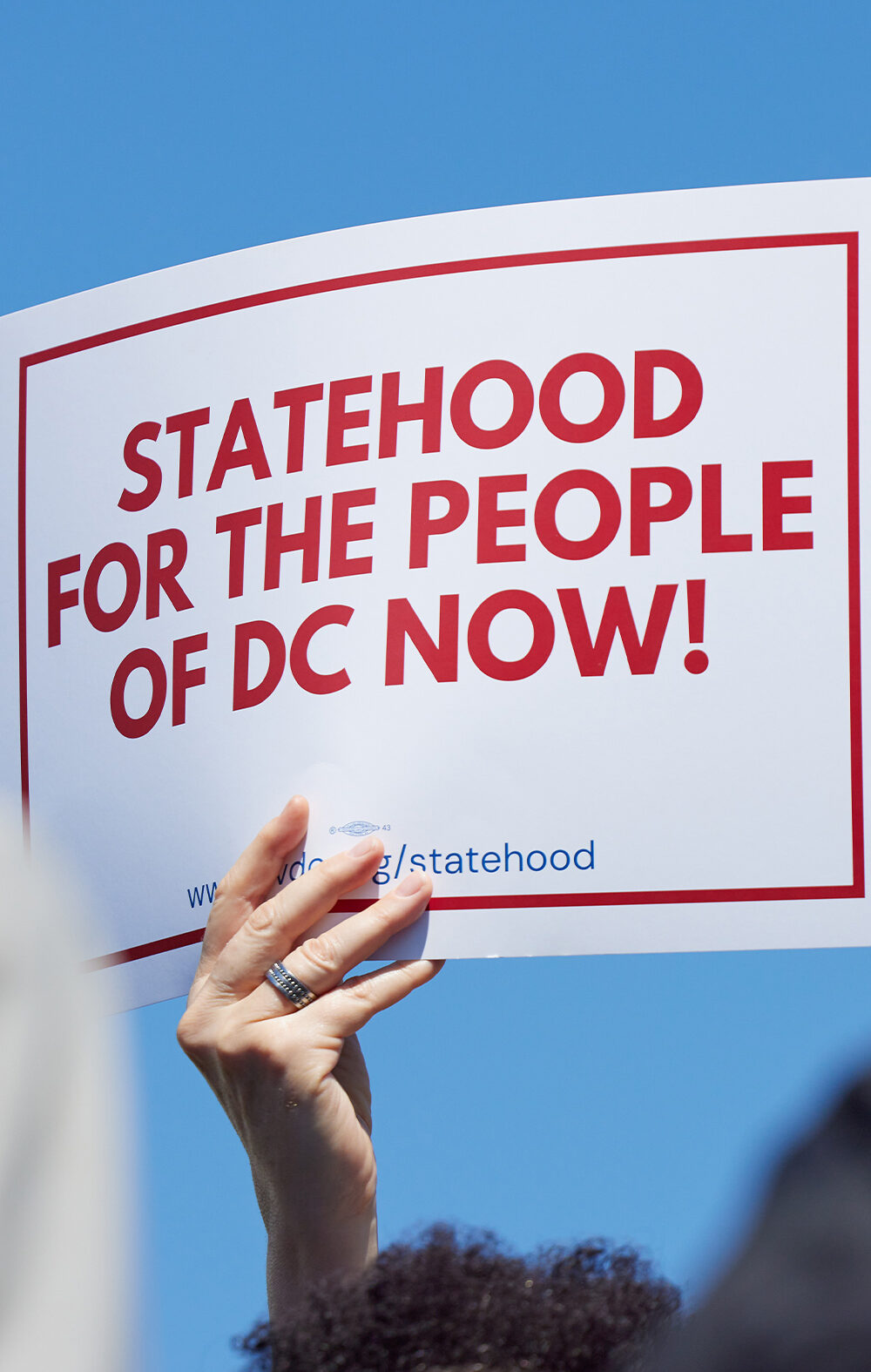 A person holds a white sign with bold red text and a red border reading "Statehood for the People of DC Now!" against a bright blue sky.