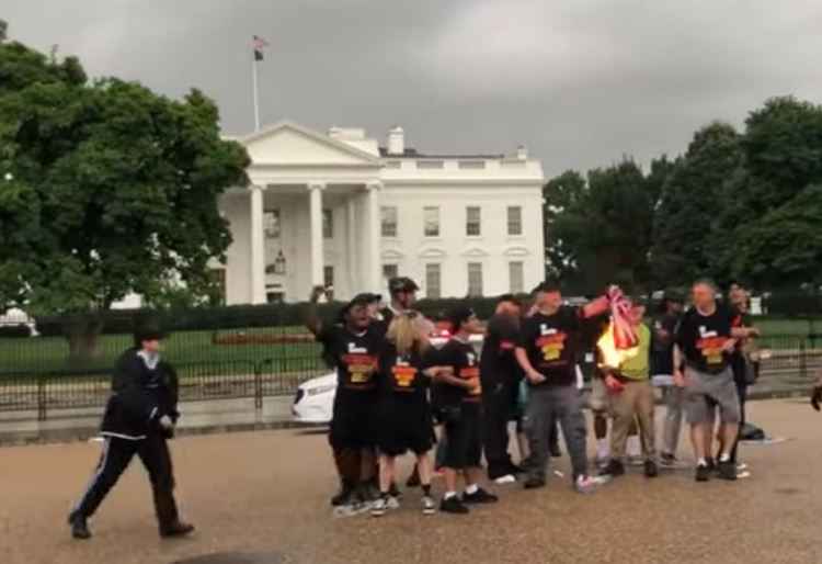 ID: a group of protesters assembled on Pennsylvania Ave with the White House in the background. Joey Johnson, a white man, has lit an American flag on fire. Flames have licked up along the fabric as US Park Police officers approach the protesters.