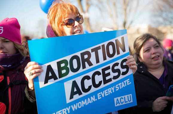 Image description: an image of a light-skinned person with red hair at a protest, holding a blue sign that states "ABORTION ACCESS: every woman, every state" with an ACLU logo in the bottom right corner of the sign