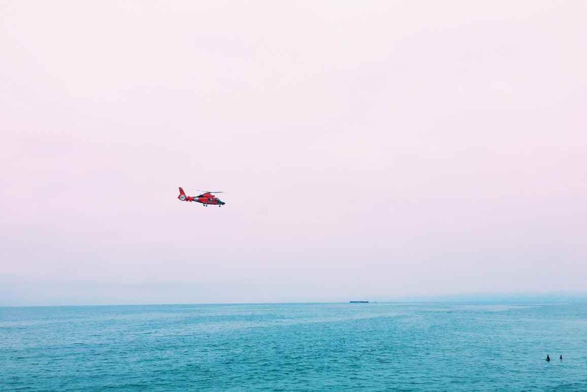 Image description: a long shot of a red helicopter above turquoise waters. Two surfers appear as small dots in the lower right corner of the frame. The background horizon is a light pastel pink.