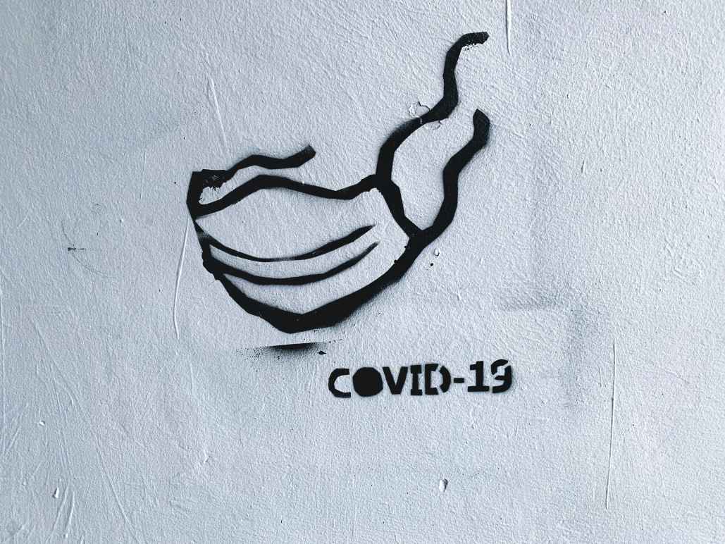 ID: a picture of a street art stencil rendered in black spray paint on a white wall. The design is of a disposable surgical mask with the ties fluttering in the wind. "COVID-19" is stenciled below. Photo by Adam Niescioruk from Unsplash.com