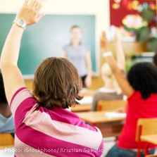 Light-skinned child raising their hand in a classroom setting with other students and a teacher in the background