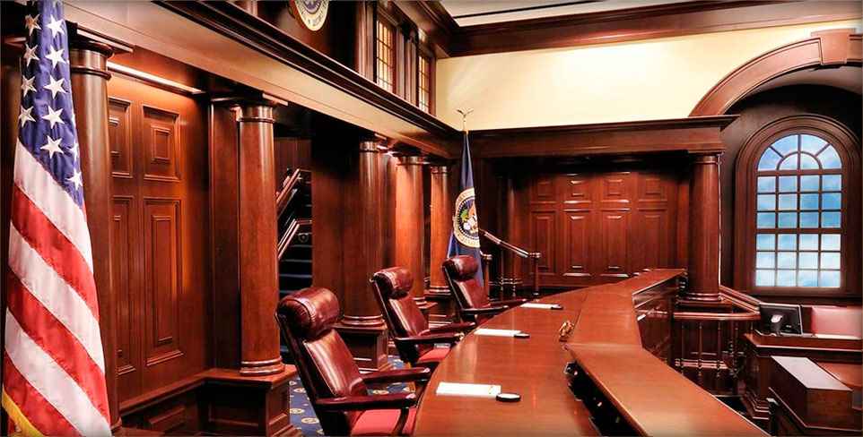 ID: the judges' bench in a courtroom in the Court of Appeals for the Federal Circuit. Three seats face to the right and the judges’ bench extends outwards into the background. Mahogany paneling glows from the light overhead.