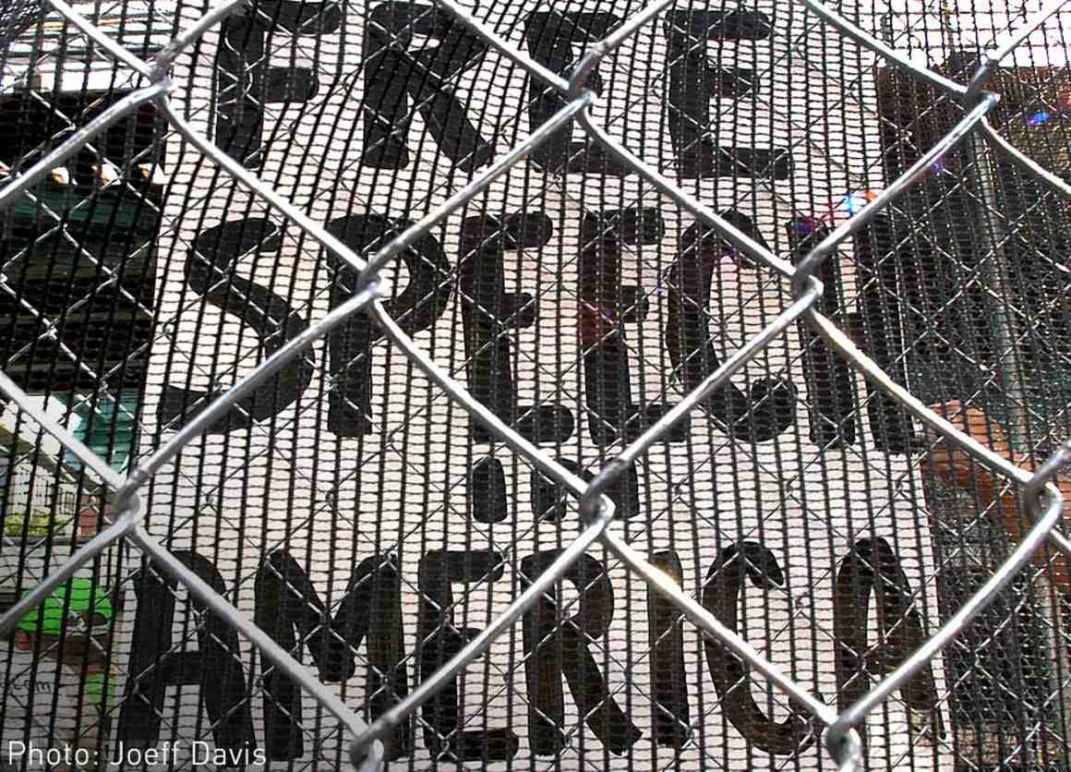 Image description: a sign stating "Free speech in America" behind a chainlink fence. 