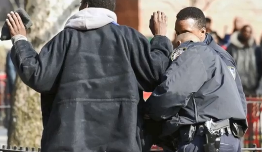 Image description: image of a person of color with his hands up and getting searched by a police officer