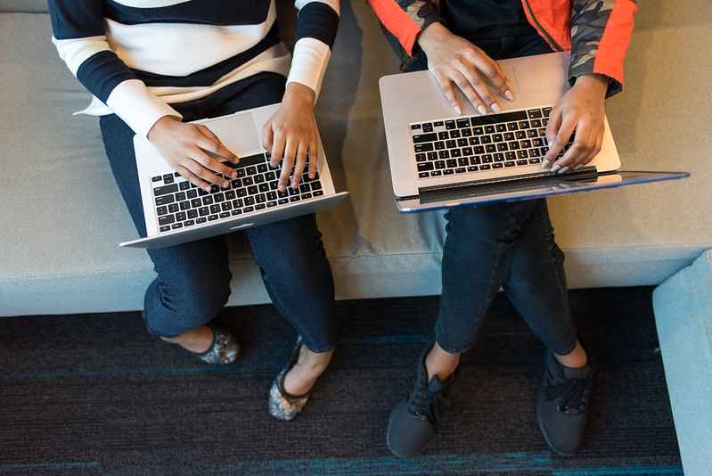 ID: overhead view of 2 computer users who have melanated skin tone laptops on their laps. User on the left has a striped sweater; they're in the middle of typing. User on the right has 1 hand on the trackpad and the other resting closer to the screen.