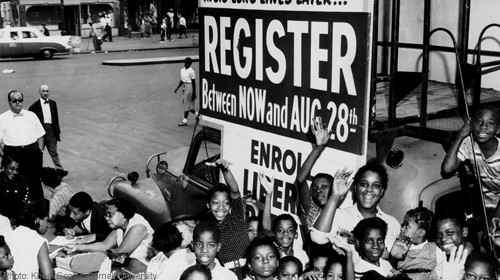 Image from the 1960's of African American youth smiling and gesturing in front of a sign that encourages black voters to register