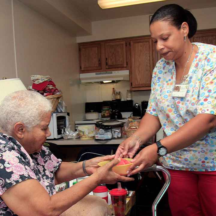 A home health care aide handing her patient a bowl of food.